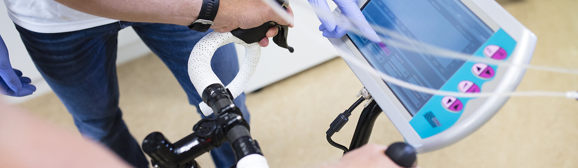 Seitliche Perspektive einer Person, die eine Leistungsdiagnostik durchführt. Ein Sportwissenschaftler betreut den Sportler, während ein Bildschirm die Werte der Diagnostik anzeigt.